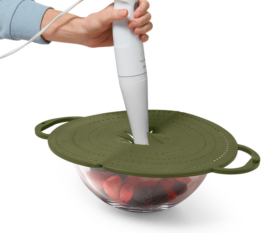 Eine Person benutzt einen Stabmixer durch den 4-in-1-Pfannenspritzschutz auf einer Schüssel mit Früchten.