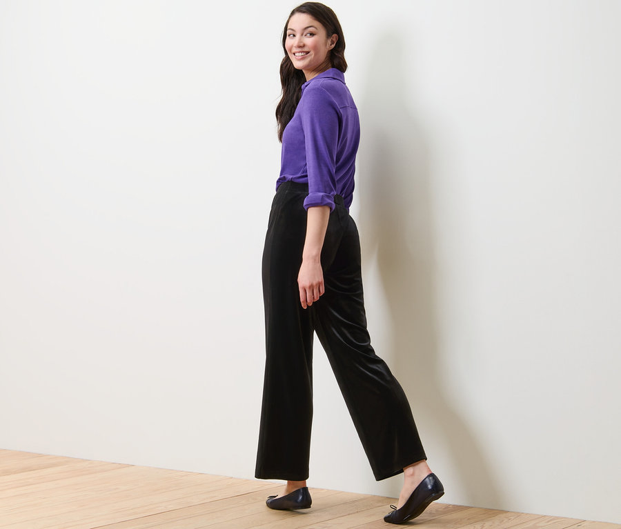 Frau geht und schaut über die Schulter, sie trägt ein lila Hemd und eine schwarze Samt-Palazzohose.