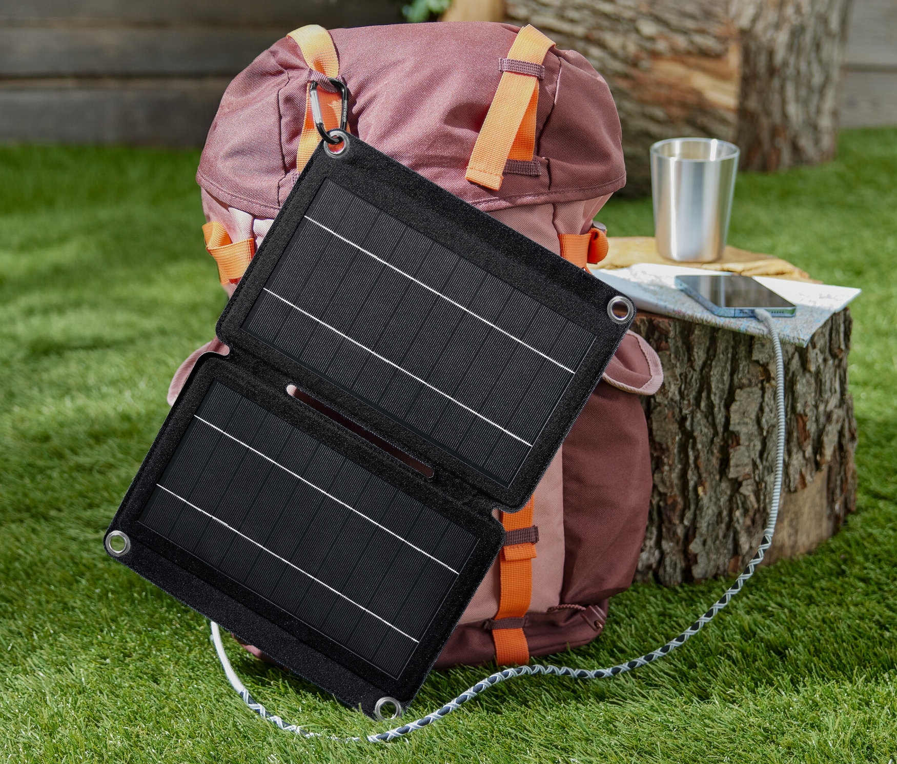 Ein faltbares Solar-Ladegerät mit 10 W hängt an einem Rucksack. Auf einem Baumstumpf wird ein Telefon geladen.