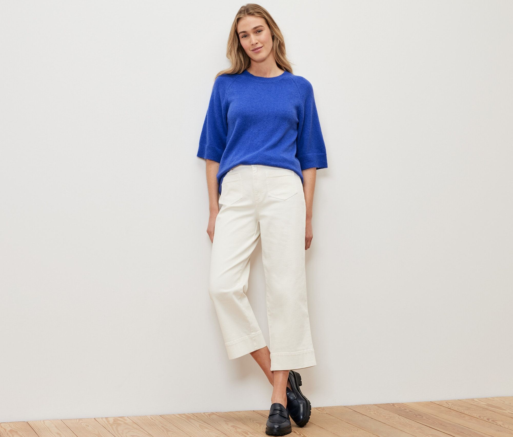 Eine Frau steht vor einer weißen Wand und trägt einen kobaltblauen Strickpullover, weiße Hosen und schwarze Schuhe.
