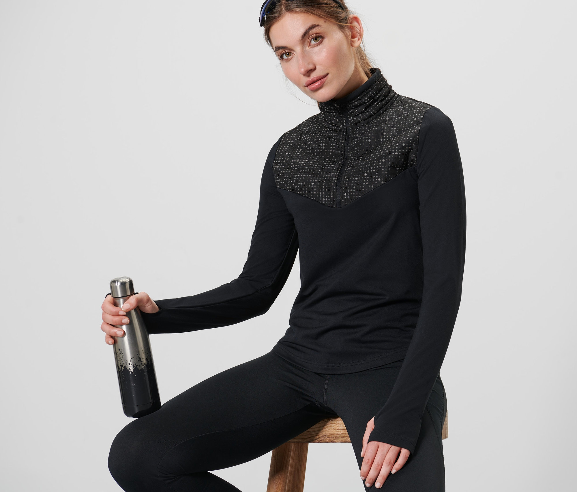 Frau sitzt auf einem Hocker und hält eine Thermosflasche. Sie trägt ein schwarzes Hybrid-Thermoshirt und schwarze Leggings.