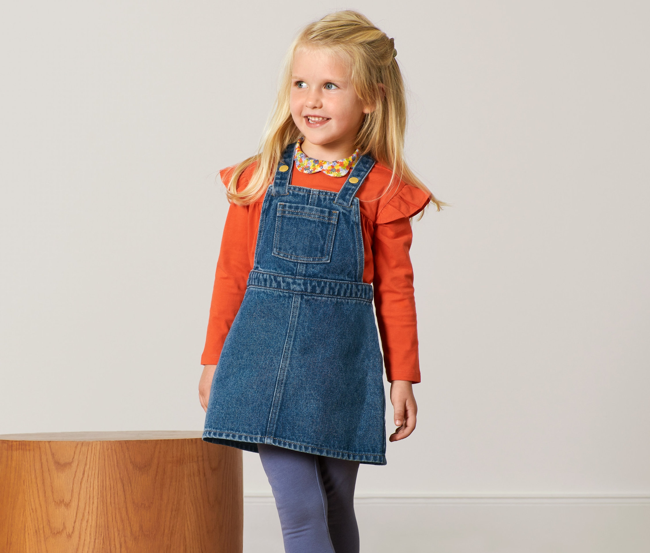 Lächelndes blondes Mädchen trägt ein Jeans-Latzkleid und ein orangefarbenes Hemd.