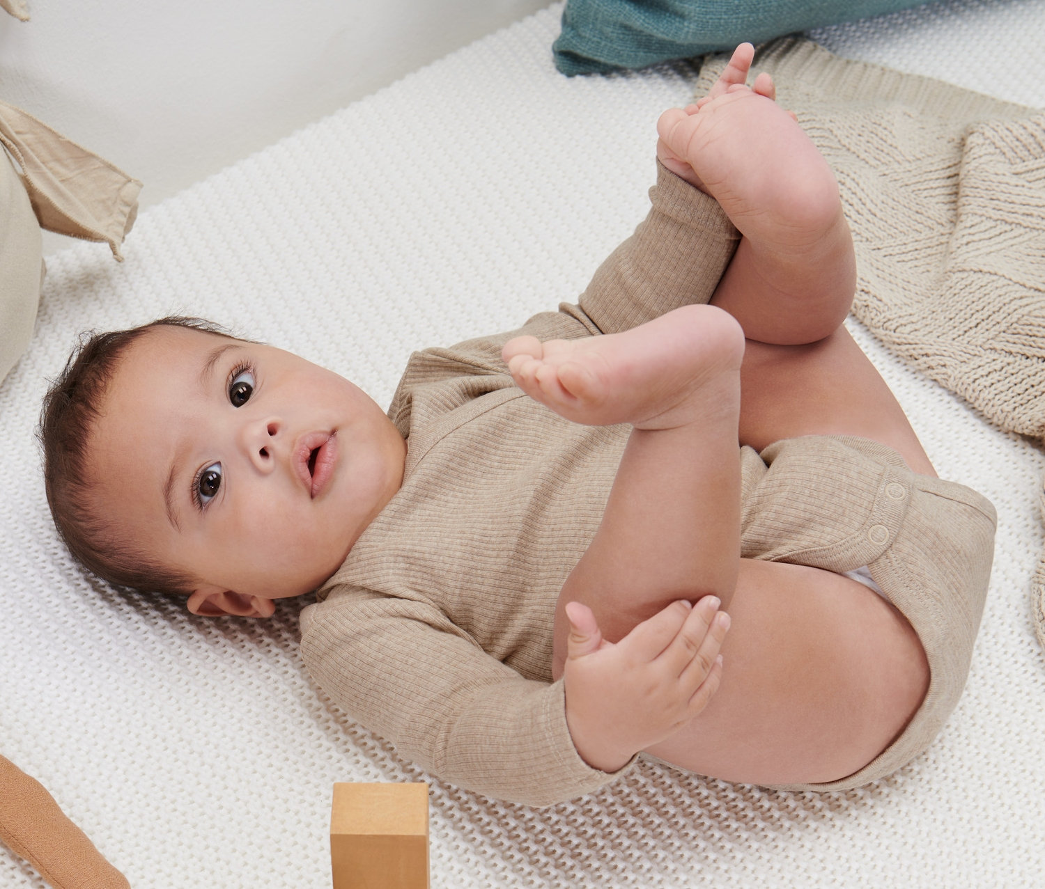 Ein Baby liegt auf dem Rücken auf einer weißen Strickdecke und spielt mit seinen Füßen. Es trägt einen beigen Langarmbody.