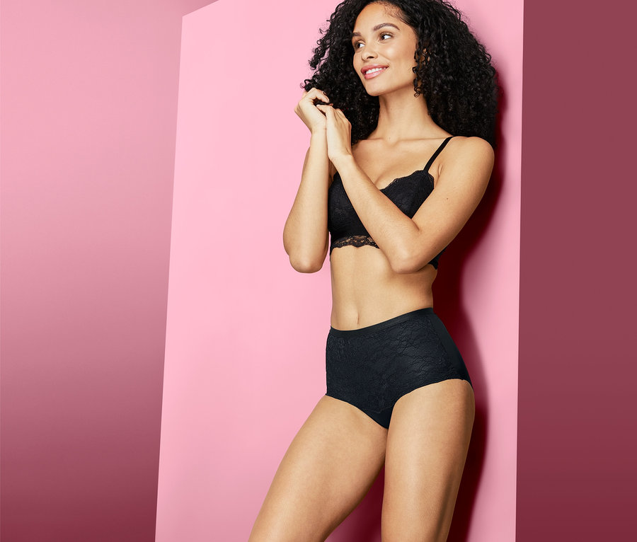 Frau mit dunklen, lockigen Haaren posiert in schwarzen Bodyforming-Panty und Spitzen-BH vor rosa Hintergrund.