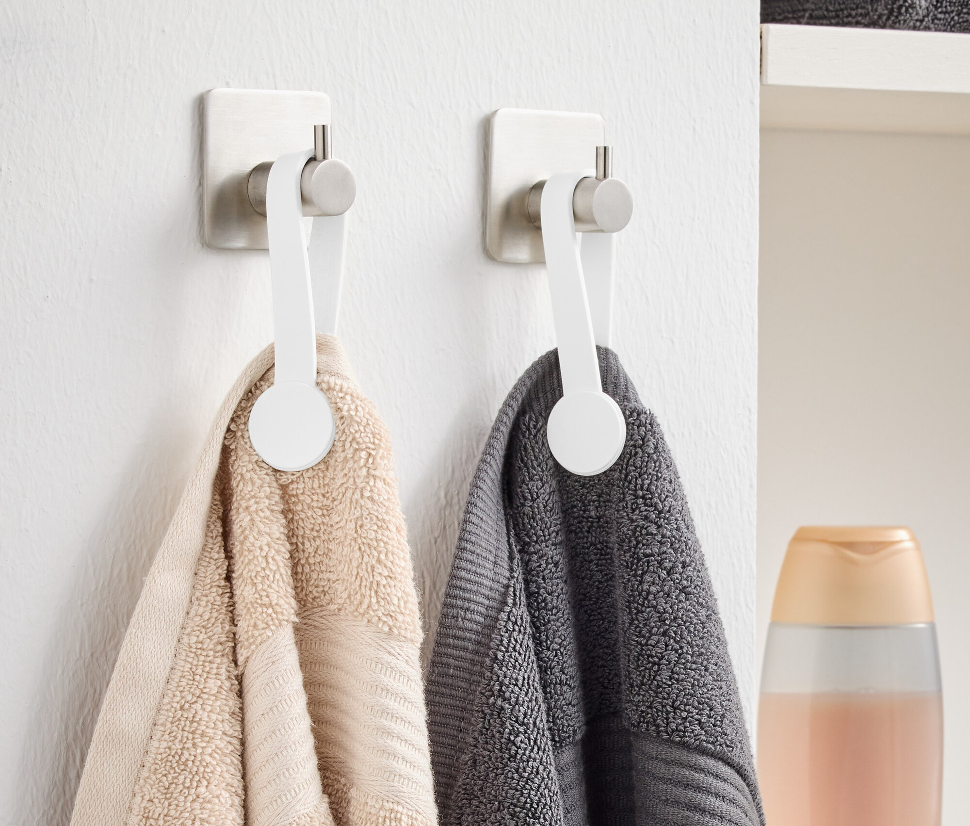 Zwei Handtücher, ein beiges und ein graues, hängen an weißen Haken an der Wand. Im Hintergrund sind gefaltete graue Handtücher und eine Flasche zu sehen.