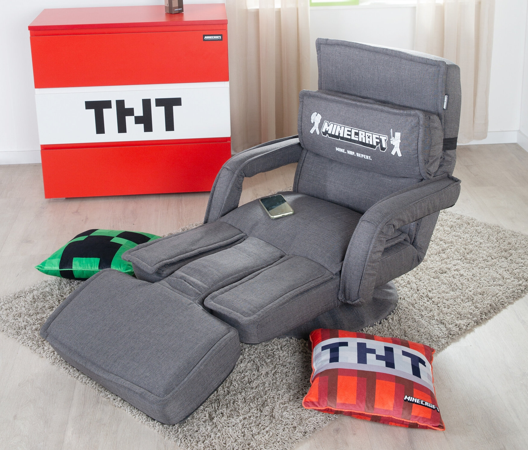 Ein grauer roba Minecraft Loungesessel mit Beinen und Armlehne steht auf einem Teppich. Daneben sind ein TNT-Schrank und zwei Minecraft-Kissen.