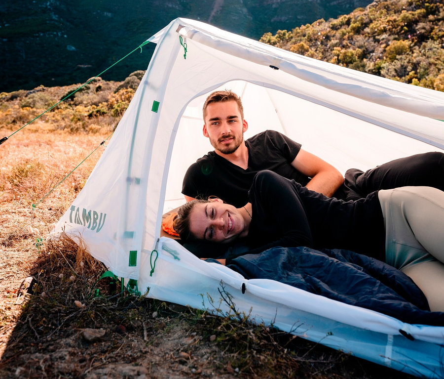 Ein lächelnder Mann und eine Frau liegen in einem weißen Tambu Suran 2-Personen-Trekkingzelt vor einem Berg.