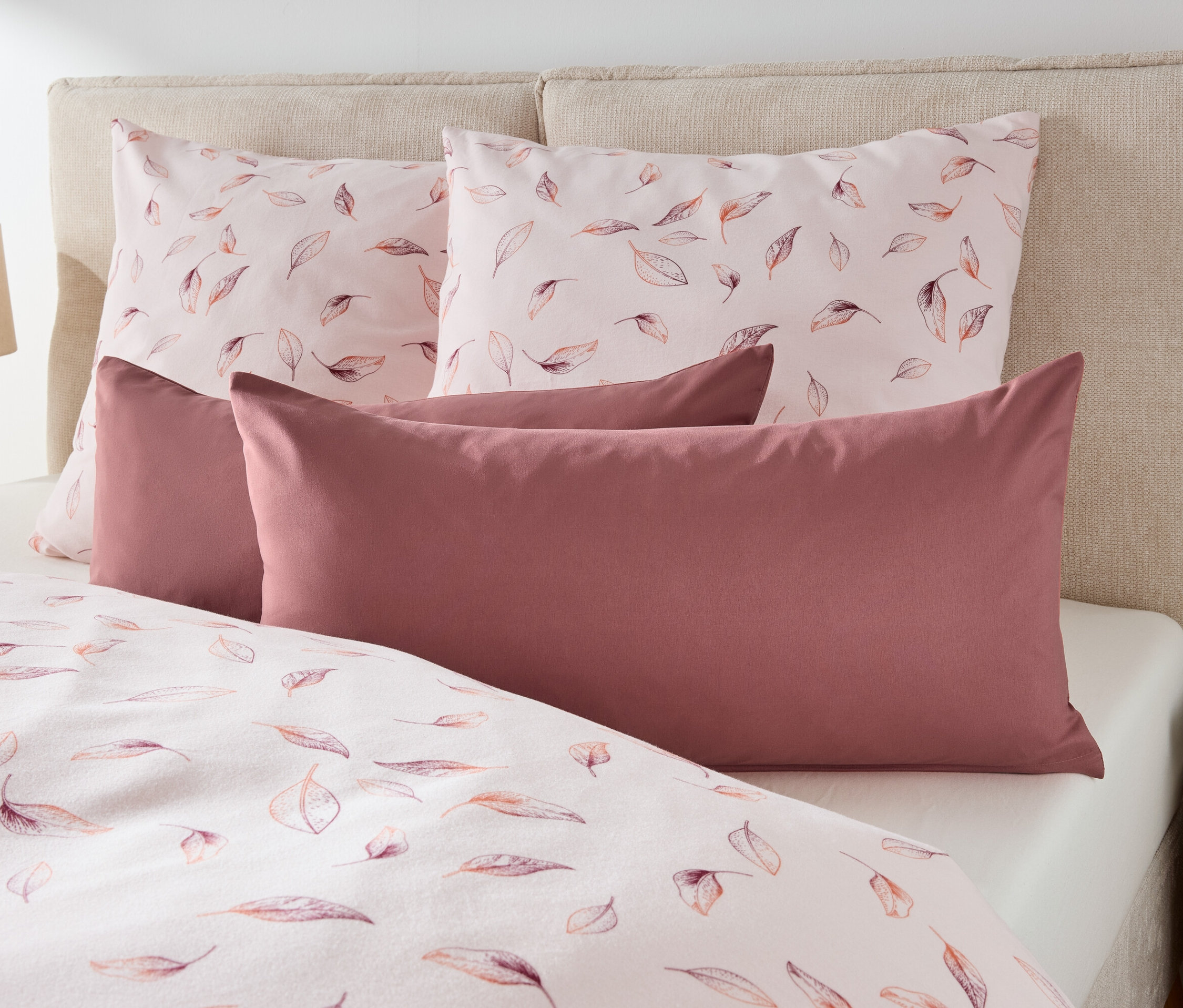 Auf einem Bett liegen zwei Kissen mit Blätterdruck und zwei kastanienbraune Kissen.