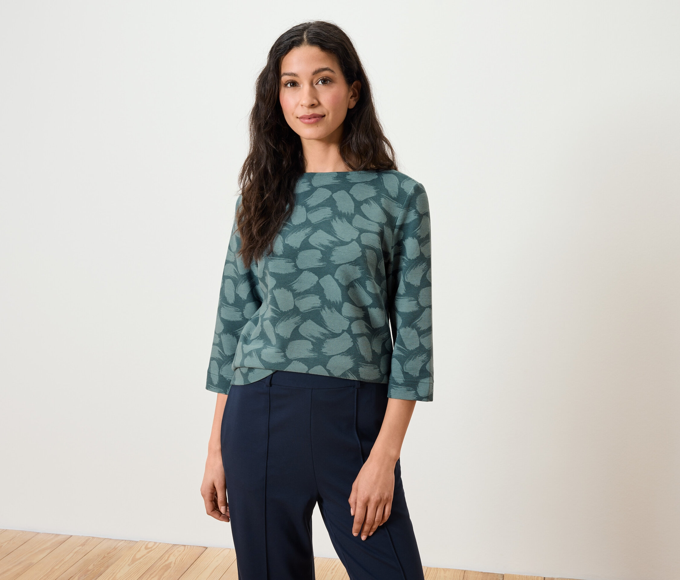 Frau mit braunen Haaren posiert in einem Oberteil mit Dreiviertelärmeln und dunkelblauen Hosen.