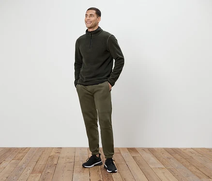 Ein Mann in olivgrünen Hosen und einem dunkelgrauen Fleece-Pullover mit Reißverschluss steht auf einem Holzboden.