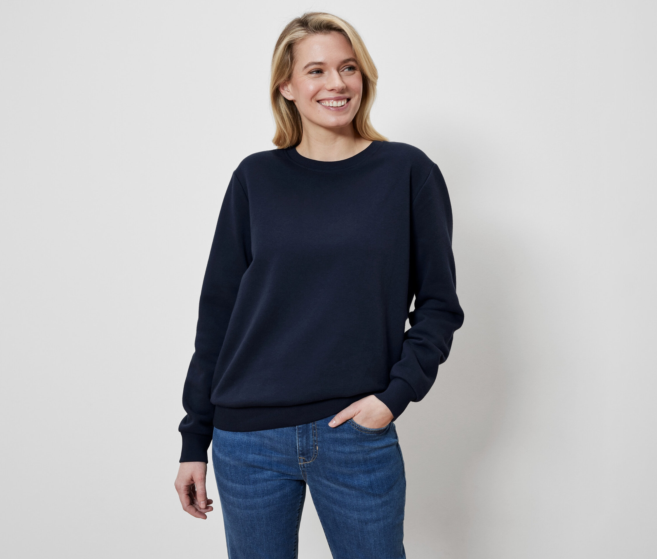 Blonde Frau mit der Hand in der Hosentasche lächelt und trägt ein dunkelblaues Sweatshirt.
