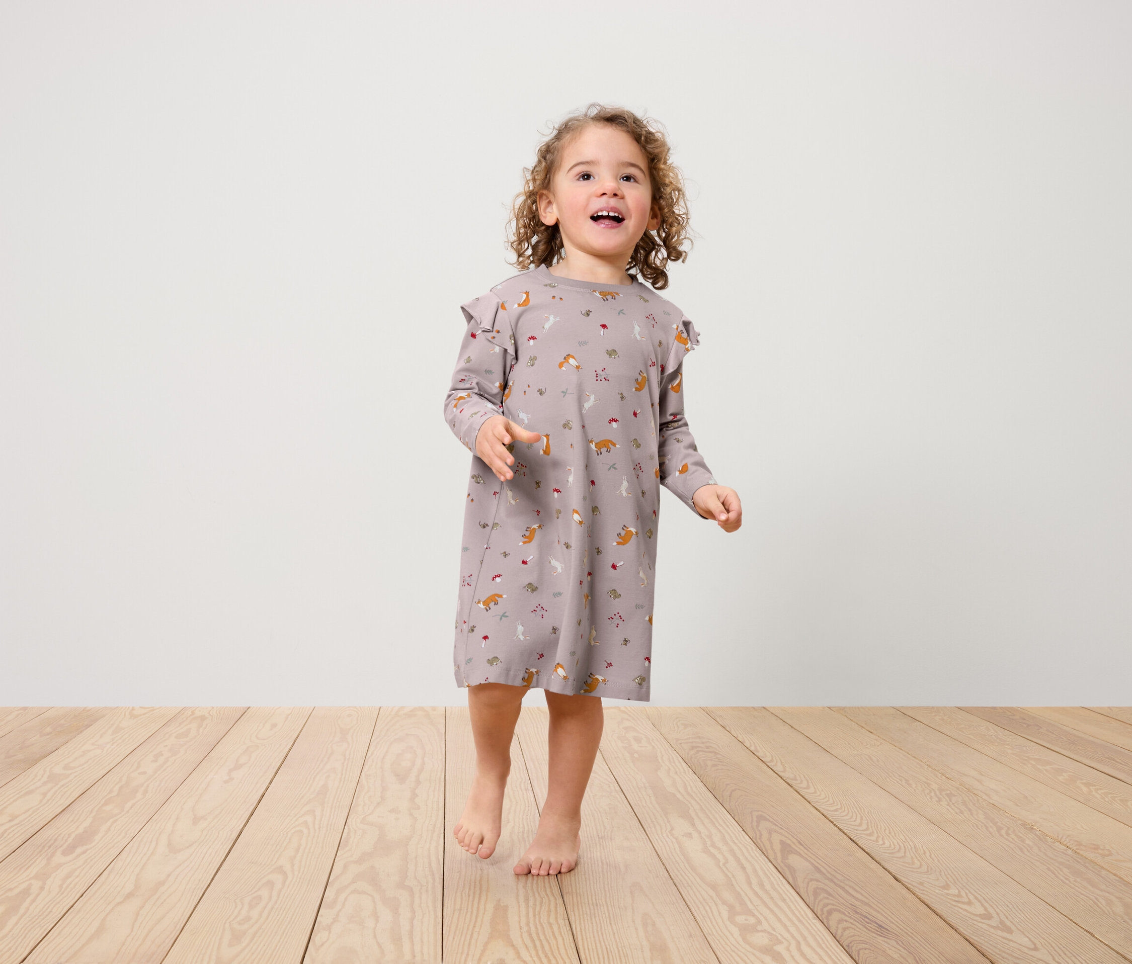 Ein kleines Mädchen in einem Kleid mit Fuchsmuster und Rüschen an den Schultern steht auf einem Holzboden.