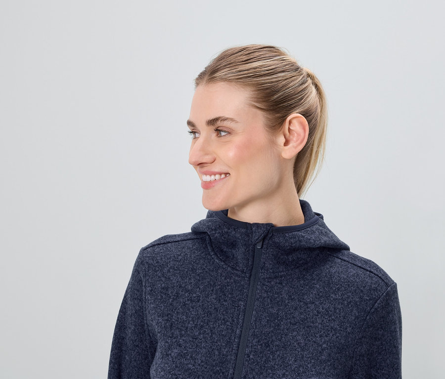 Profilansicht einer lächelnden Frau mit blonden Haaren in einer blauen Reissverschluss-Kapuzenjacke.