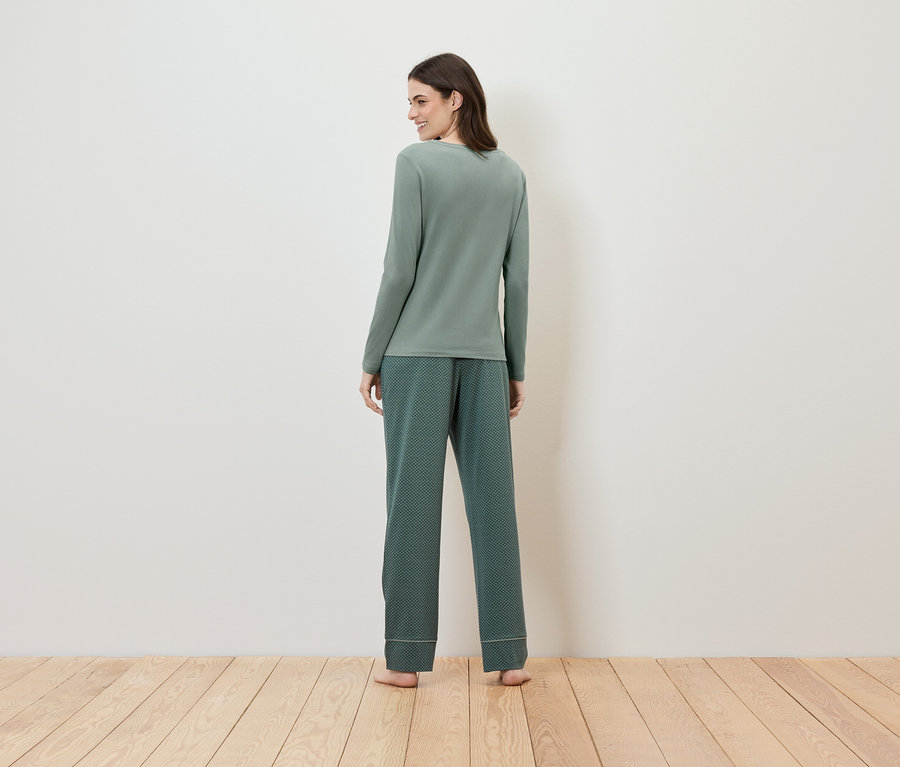 Frau in grünem langärmligem Pyjama mit gemusterter Hose steht mit dem Rücken zur Kamera.