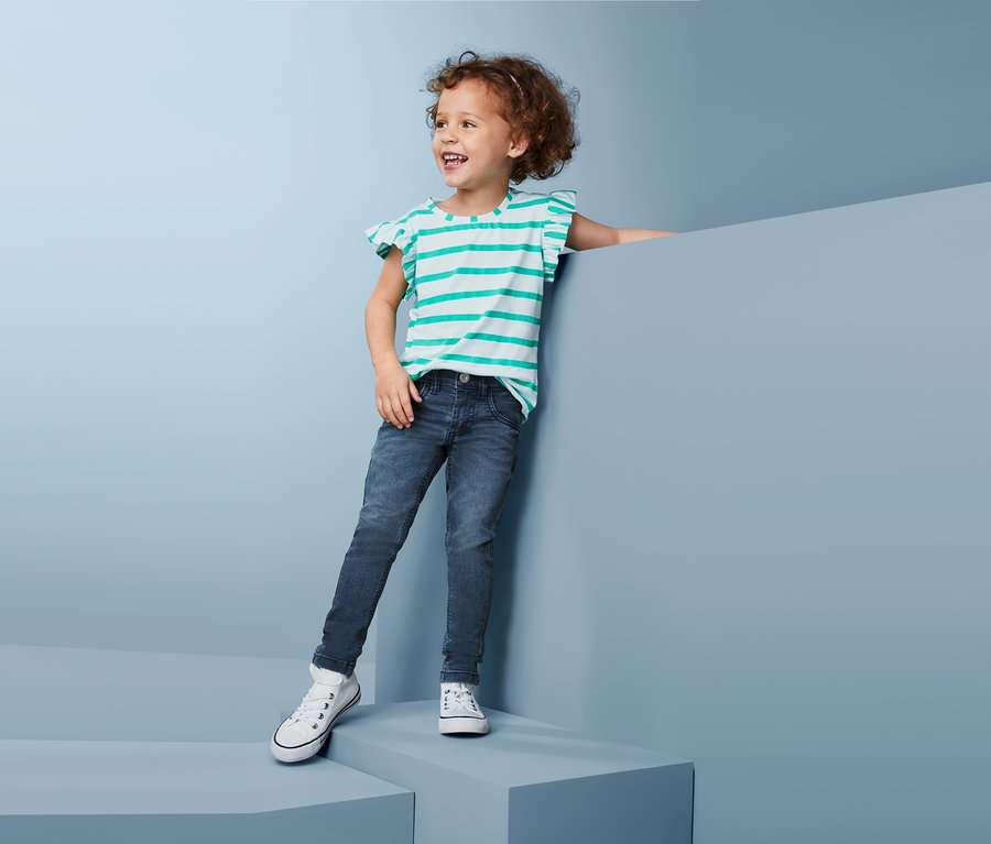 Ein lächelndes kleines Mädchen steht da und lehnt sich an eine blaue Wand. Sie trägt blaue Slimfit-Stretchdenim und ein gestreiftes T-Shirt.
