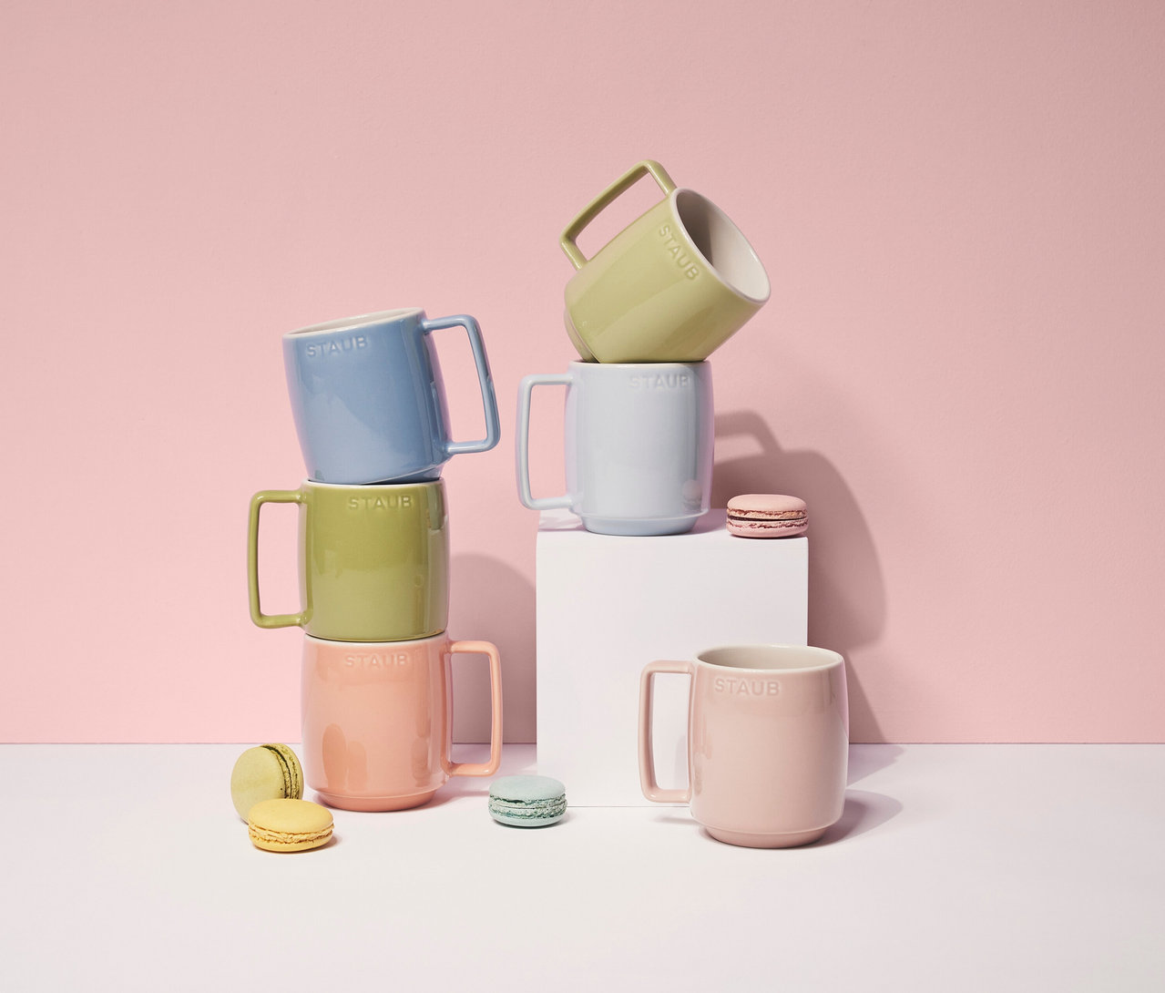 Sechs STAUB Kaffeebecher »Macaron« in Pastellfarben sind gestapelt und auf einer weißen Oberfläche platziert, zusammen mit farblich passenden Macarons.