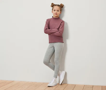 Ein Mädchen mit braunen, zu Duttfrisuren hochgesteckten Haaren steht mit verschränkten Armen vor einer Wand. Sie trägt ein weinrotes Langarmshirt, graue Leggings und weiße Turnschuhe.
