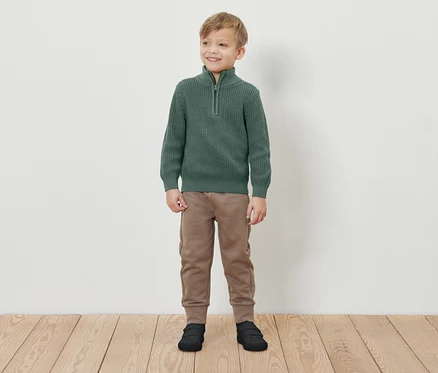 Ein Junge steht lächelnd auf einem Holzboden und trägt einen grünen Pullover mit Reißverschluss, braune Jogginghose und schwarze Schuhe.