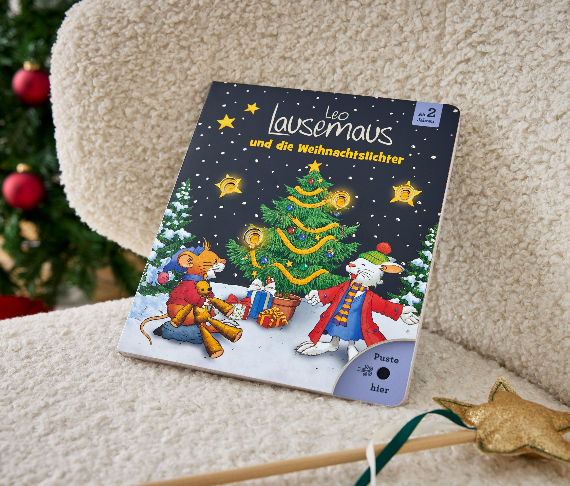 Kinderbuch Leo Lausemaus und die Weihnachtslichter liegt auf einem cremefarbenen Sessel neben einem Weihnachtsbaum.