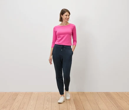 Frau mit pinkfarbenem Oberteil und marineblauer Hose steht auf einem Holzboden vor einer weißen Wand.