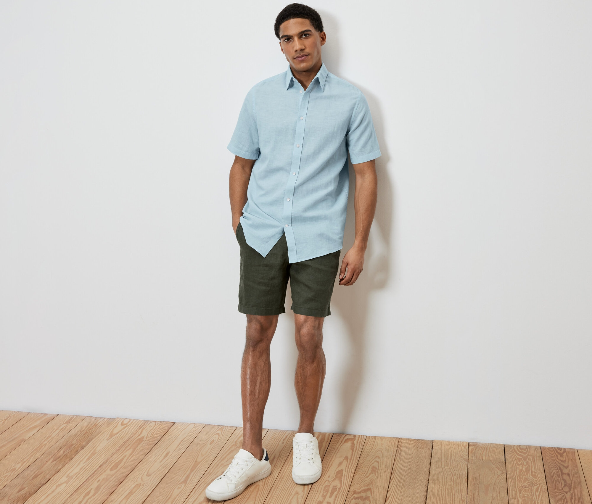 Ein Mann posiert auf dem Foto und trägt ein hellblaues Kurzarmhemd mit Leinen, Khaki-Shorts und weiße Turnschuhe.