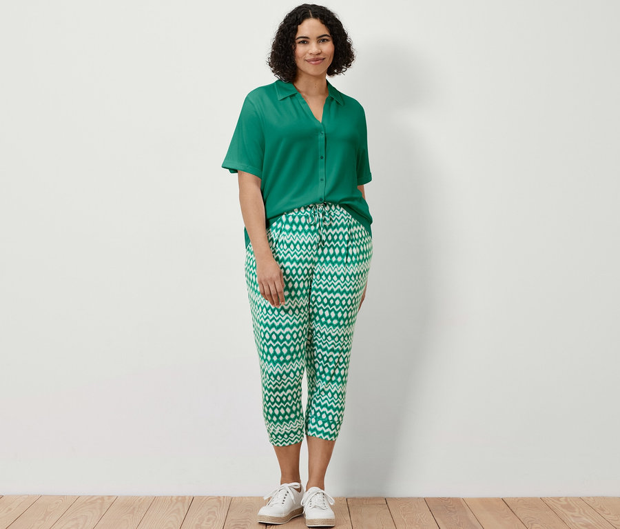 Eine Frau steht und trägt eine smaragdgrüne Blusenshirt mit Knopfleiste und eine bedruckte 3/4-Jerseyhose.