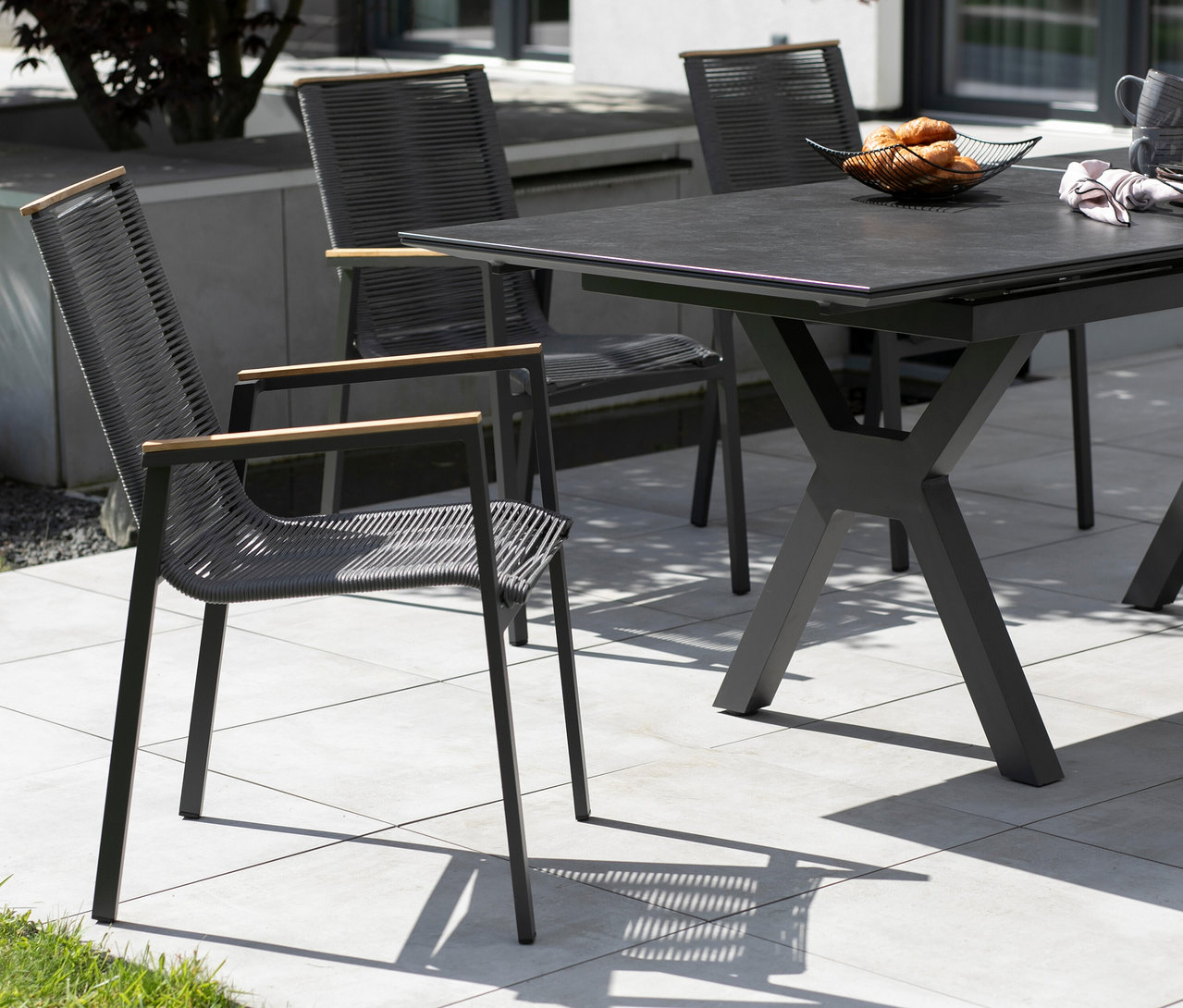 Drei graue Stühle mit Armlehnen aus Holz stehen um einen dunklen Tisch mit Gebäck im Freien.