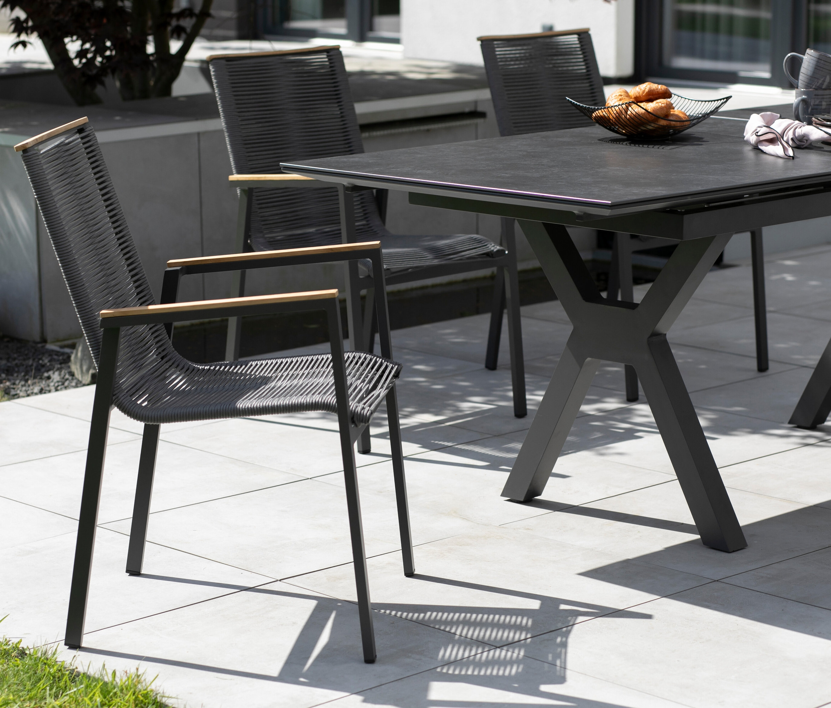 Drei graue Stühle mit Armlehnen aus Holz stehen um einen dunklen Tisch mit Gebäck im Freien.