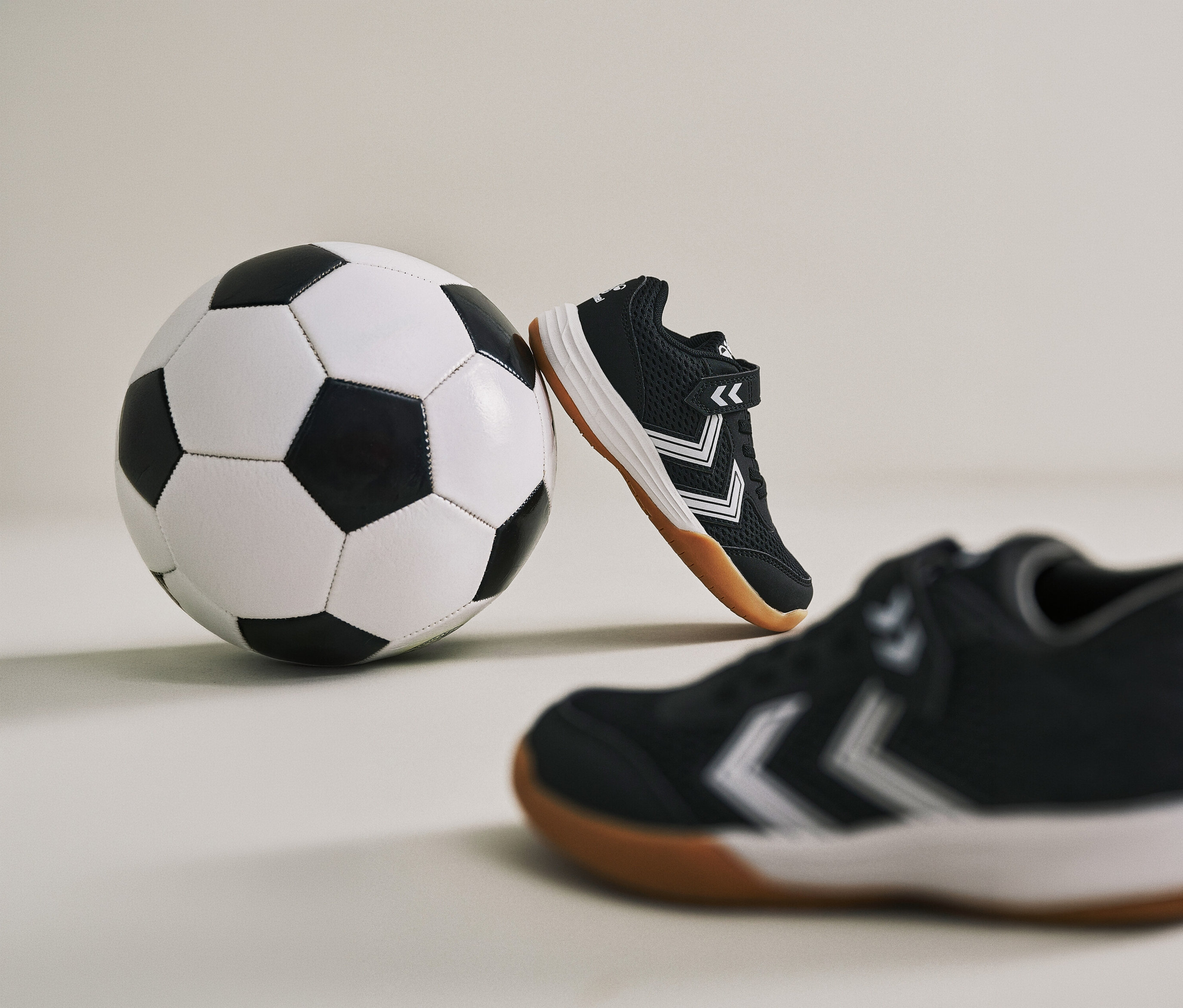 Ein Fußball lehnt an einem schwarzen hummel® Multiplay Flex VC JR Schuh. Ein weiterer Schuh ist im Vordergrund zu sehen.