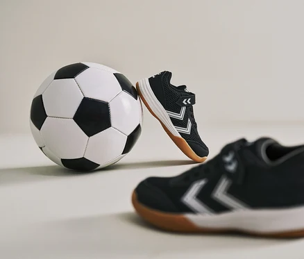 Ein Fußball lehnt an einem schwarzen hummel® Multiplay Flex VC JR Schuh. Ein weiterer Schuh ist im Vordergrund zu sehen.