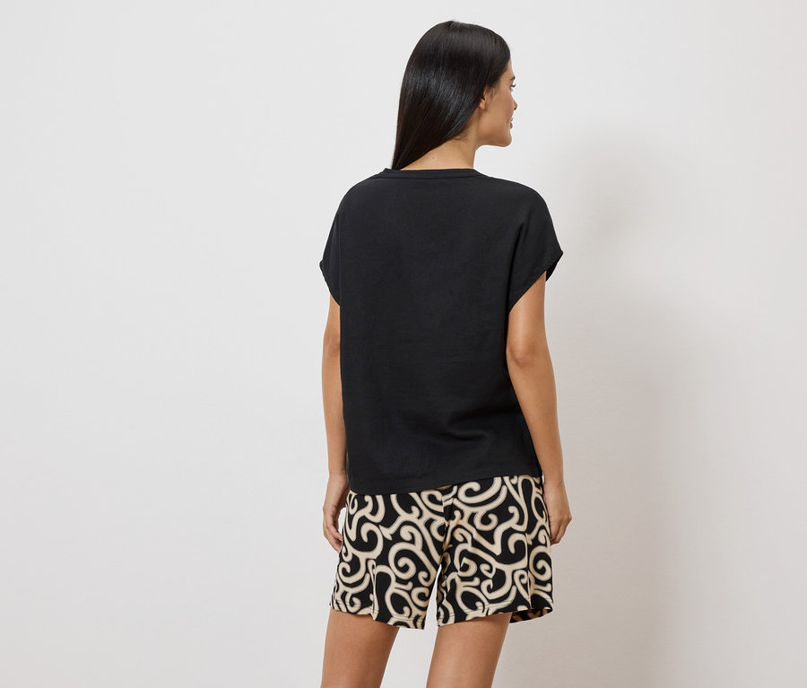 Frau von hinten in schwarzem T-Shirt und gemusterten Shorts vor weißem Hintergrund.