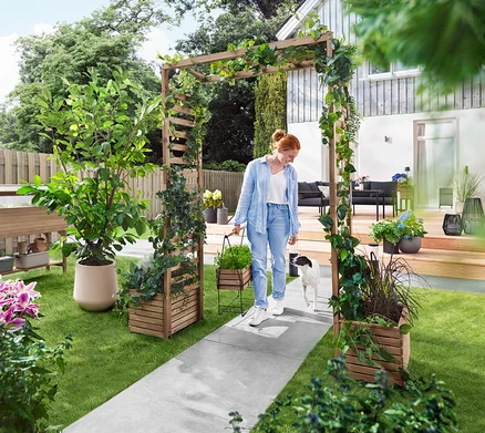 Eine Frau geht mit einem Hund durch einen Gartenbogen, der mit Pflanzen bewachsen ist. Sie trägt eine Pflanzbox.
