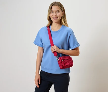 Eine Frau mit blonden Haaren lächelt und trägt ein hellblaues T-Shirt und eine rote Umhängetasche.