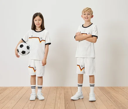 Ein Mädchen hält einen Fußball, ein Junge steht daneben, beide tragen passende weiße Trikots und Shorts mit deutschen Farben.