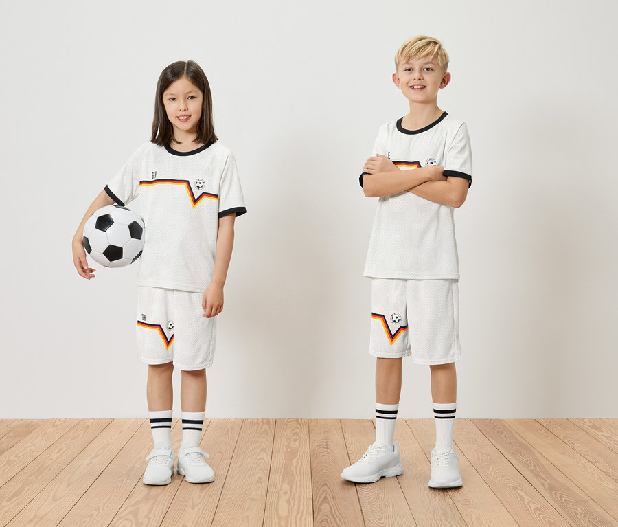 Ein Mädchen hält einen Fußball, ein Junge steht daneben, beide tragen passende weiße Trikots und Shorts mit deutschen Farben.
