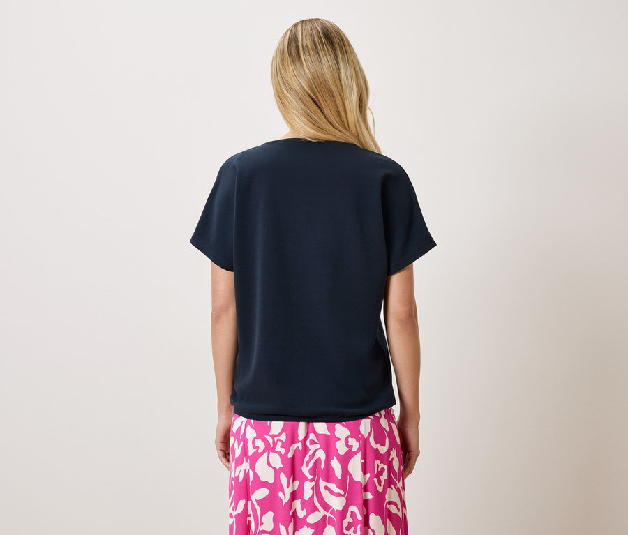 Rückenansicht einer Frau mit blonden Haaren, die ein marineblaues T-Shirt und einen pinkfarbenen Rock mit Blumendruck trägt.