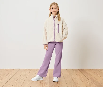 Mädchen mit beiger Teddyjacke mit lila Details, lila Hose und weißen Turnschuhen steht vor einer weißen Wand.