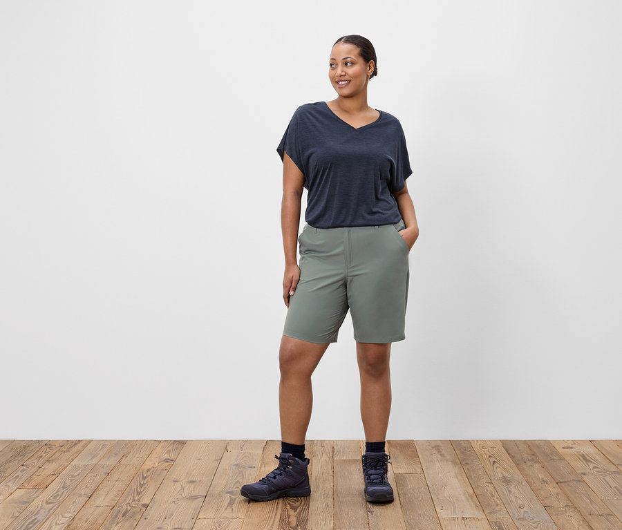 Eine Frau steht mit den Händen in den Hosentaschen, trägt ein marineblaues T-Shirt, grüne Shorts und Wanderschuhe.