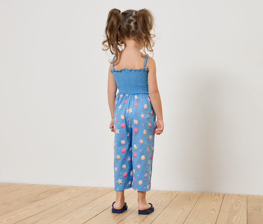 Rückansicht eines kleinen Mädchens mit Zöpfen, das einen blauen Jumpsuit mit Meerestier-Motiv und blaue Sandalen trägt.