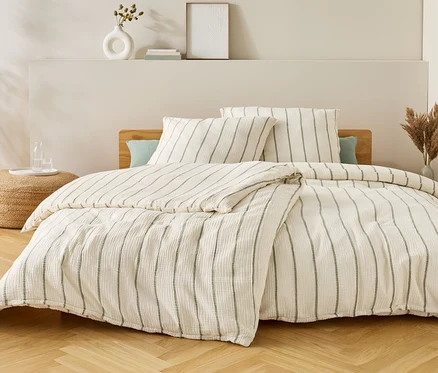 Bett mit gestreifter Bettwäsche, zwei Kissen und einer Decke. Auf dem Nachttisch stehen eine Vase und Gläser mit Wasser.
