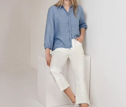 Frau steht, trägt eine hellblaue Street One Jeansbluse und eine weiße Street One Jeans mit Lochstickerei, lehnt an einer weißen Wand und einem Würfel.