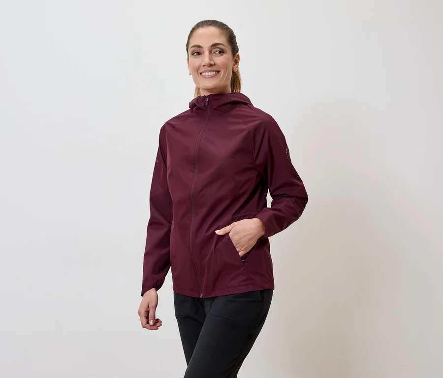 Frau mit Pferdeschwanz trägt weinrote Jacke mit Kapuze und schwarze Hose, Hand in der Jackentasche.
