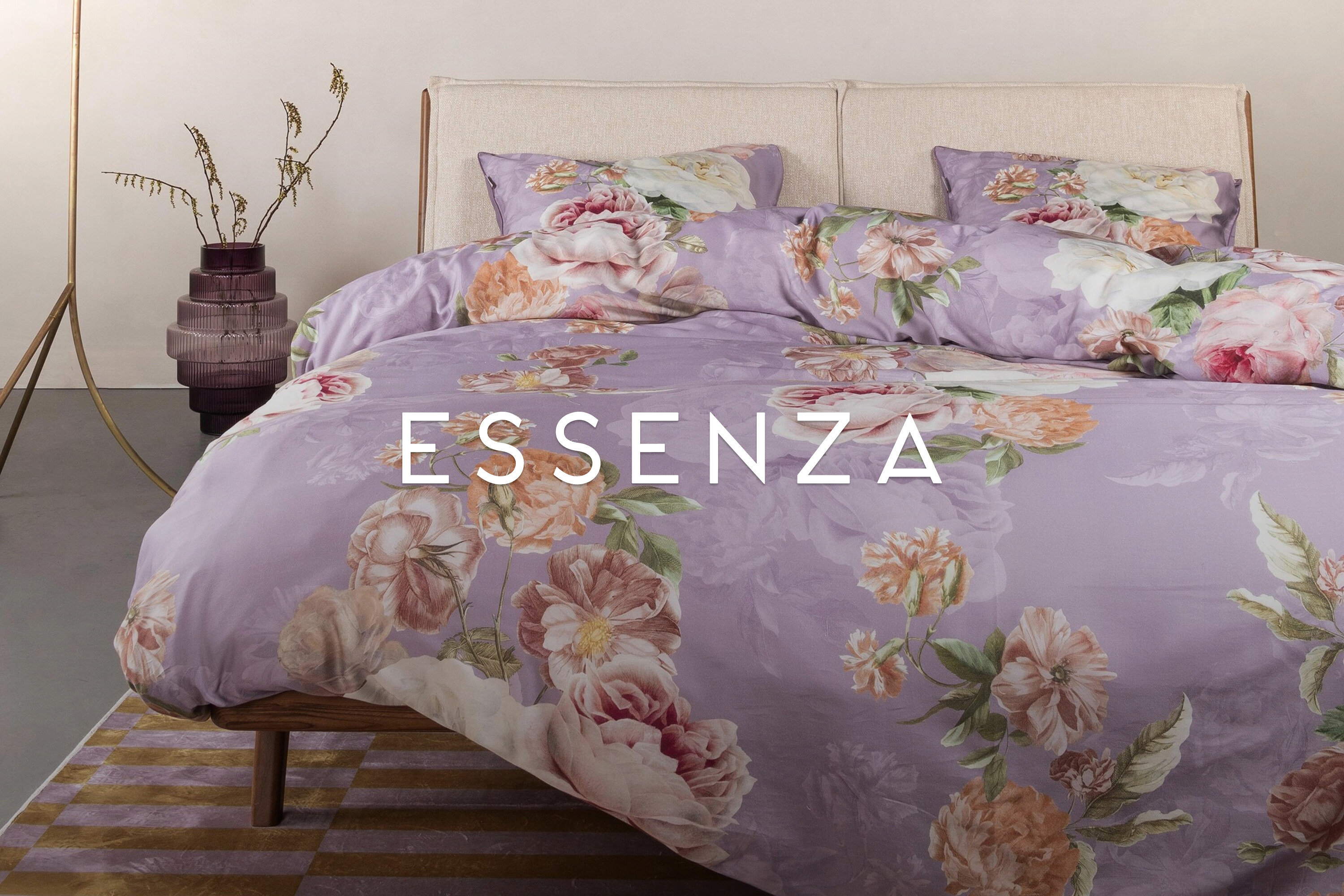 Lila Bettwäsche mit Blumenmuster liegt auf einem Bett mit Kopfteil. Eine lila Vase mit Zweigen steht links.