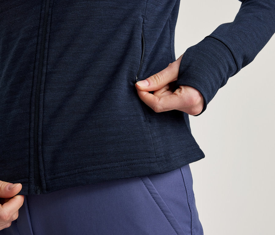 Person mit marineblauer Jacke mit Reißverschluss und Daumenlöchern, die eine Seitentasche öffnet, dazu blaue Hose.