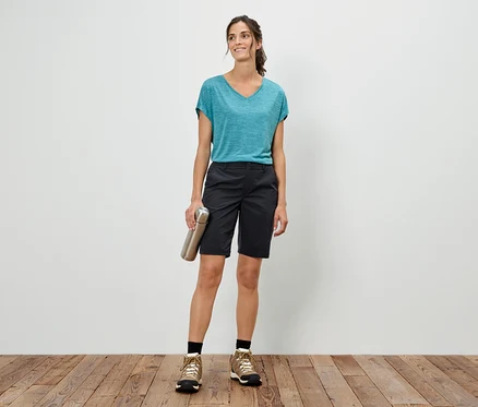 Frau steht vor einer weißen Wand, hält eine silberne Flasche, trägt ein blaues T-Shirt, Shorts und Wanderschuhe.