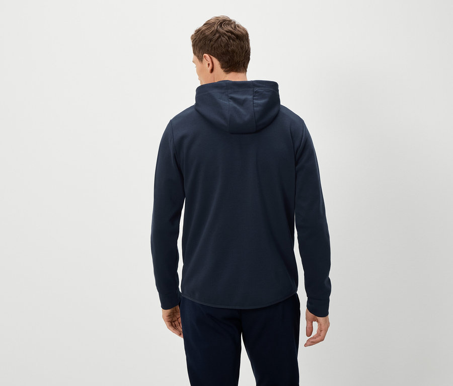 Rückansicht eines Mannes mit marineblauem Kapuzenpullover und marineblauer Hose vor einem hellen Hintergrund.