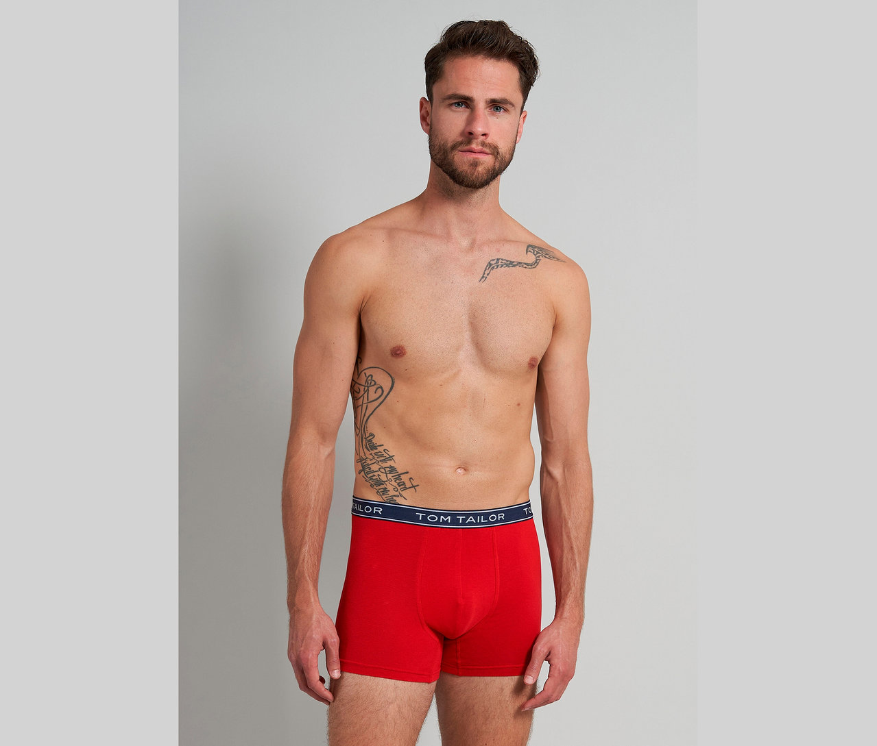 Mann mit Tätowierungen trägt rote Tom Tailor Boxershorts vor grauem Hintergrund.