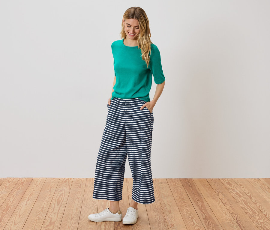 Frau steht mit Händen in den Taschen einer blau-weiß gestreiften Culotte, trägt ein grünes Oberteil und weiße Turnschuhe.