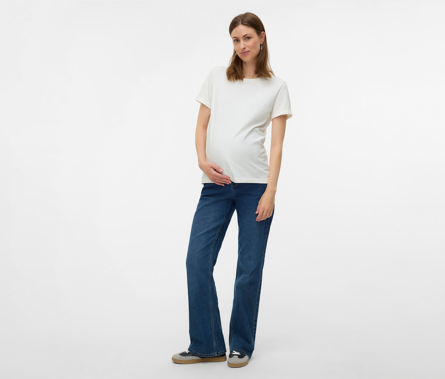 Schwangere Frau in weißem T-Shirt und MAMALICIOUS Flared Jeans »MLEDINA« posiert vor einem weißen Hintergrund.