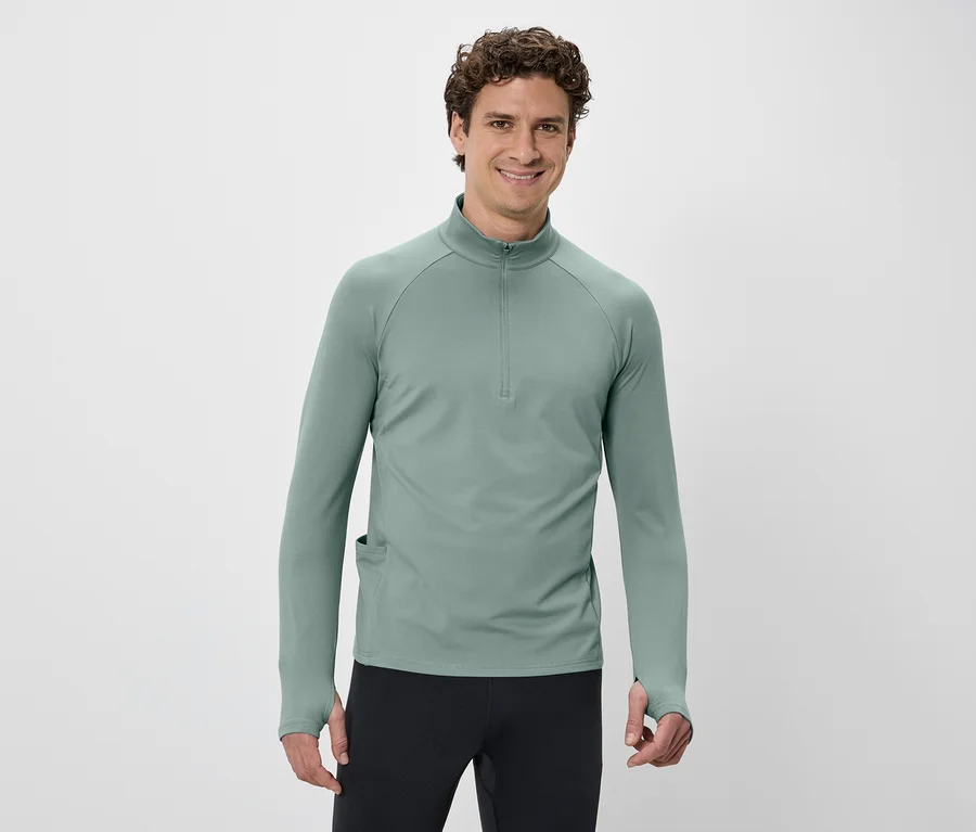 Mann mit lockigem Haar trägt ein grünes Langarmshirt mit Reißverschluss und schwarze Hose vor einem weißen Hintergrund.
